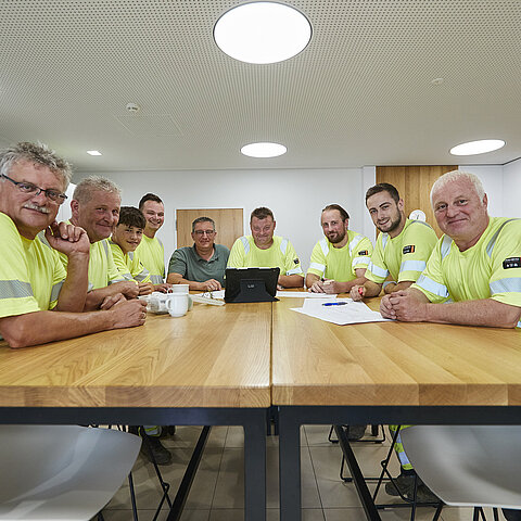 Neun Beschäftigte in Warnschutzkleidung an einem langen Tisch im Aufenthaltsraum