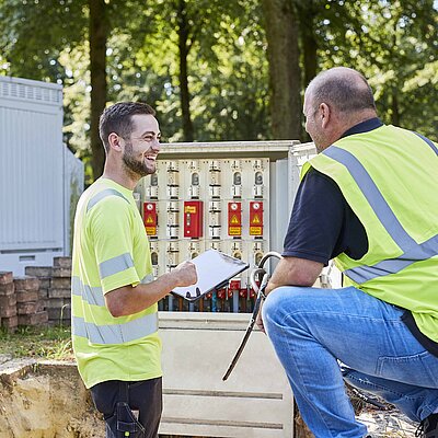 Zwei Beschäftigte im Gespräch vor einem geöffneten Verteilerschrank auf einer Baustelle