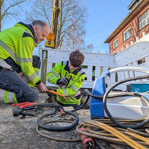Zwei Beschäftigte in Warnschutzkleidung arbeiten im Freien an Kabeln und Werkzeug