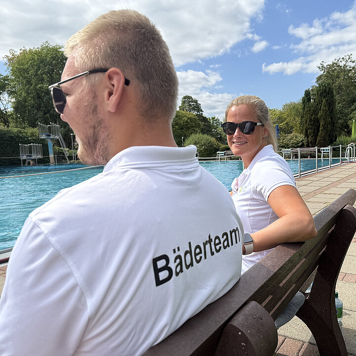 Ein Kollege und eine Kollegin sind im Freibad in der Aufsicht am Beckenrand. Der Kollege ist von hinten zu sehen, währen die Kollegin mit Sonnenbrille in die Kamera lächelt.